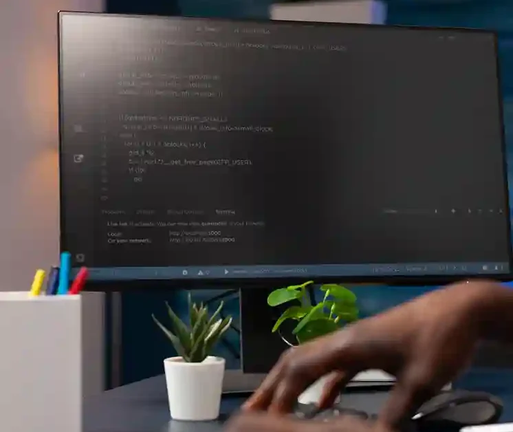 Person working at a desk using a computer monitor displaying code on a dark-themed screen in a modern office workspace.